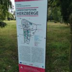 Informationsschild im Landschaftspark Herzberge, mit Karte und Beschreibung des Parks, umgeben von Bäumen und Grasflächen