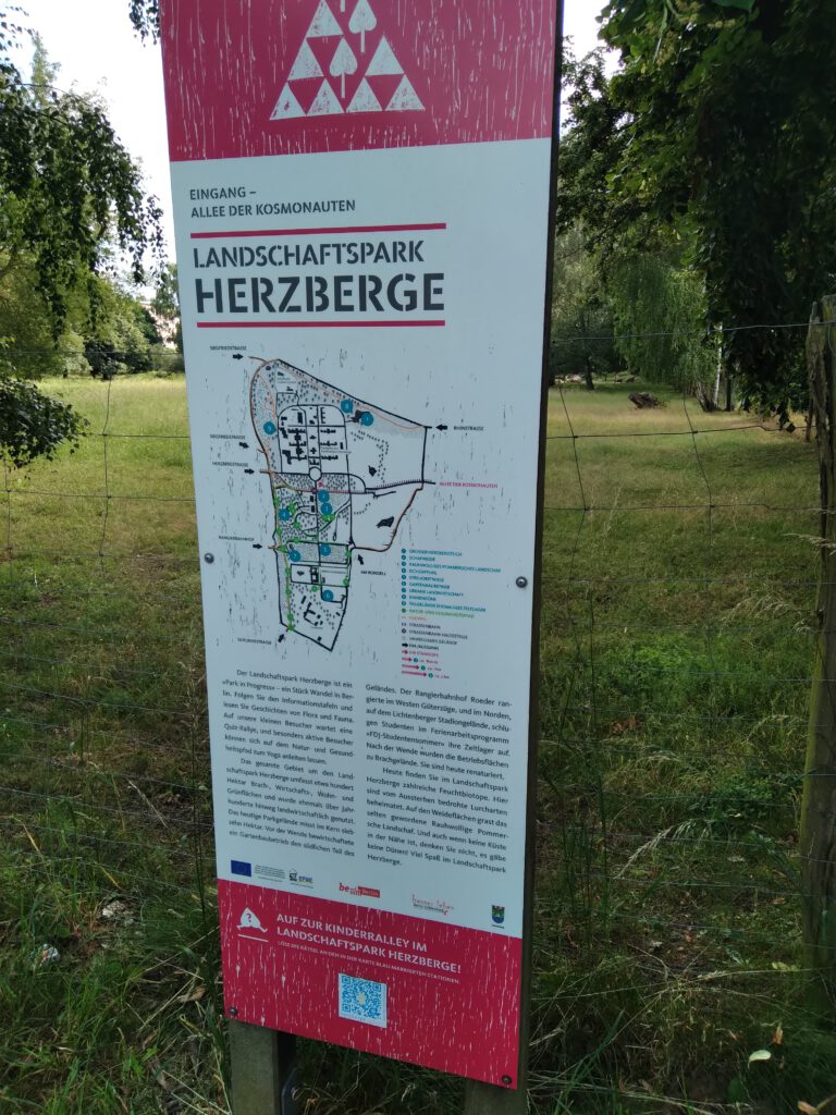 Informationsschild im Landschaftspark Herzberge, mit Karte und Beschreibung des Parks, umgeben von Bäumen und Grasflächen