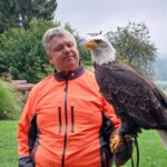 Person (Bernd Spindler) in orangefarbener Jacke steht mit einem majestätischen Greifvogel auf dem Arm bei einem Leadership-Workshop in der Stauferfalknerei Lorch.