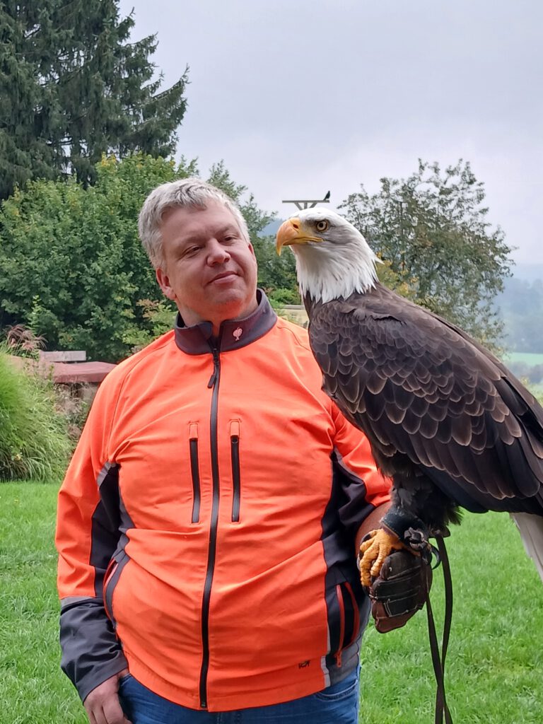 Person (Bernd Spindler) in orangefarbener Jacke steht mit einem majestätischen Greifvogel auf dem Arm bei einem Leadership-Workshop in der Stauferfalknerei Lorch.