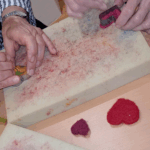 Hände beim Trockenfilzen mit Filznadeln, roter gefärbter Schafwolle und Ausstechformen auf Filzunterlage.