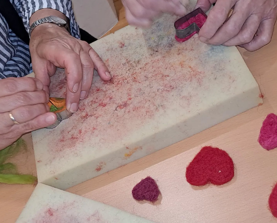 Hände beim Trockenfilzen mit Filznadeln, roter gefärbter Schafwolle und Ausstechformen auf Filzunterlage.
